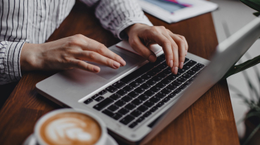 Female hands press laptop keys while sitting at wooden table with white cappuccino cup