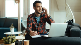 Young smiling man writing notes while making a phone call and using laptop at home.